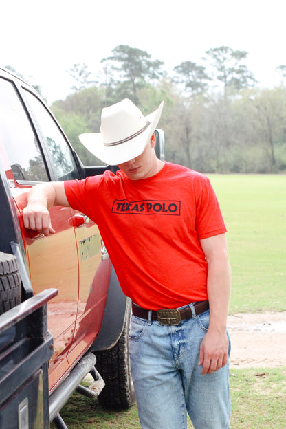 Man wearing a red t-shirt with black Texas Polo logo and tan cowboy hat, leaning against a red pickup truck.
