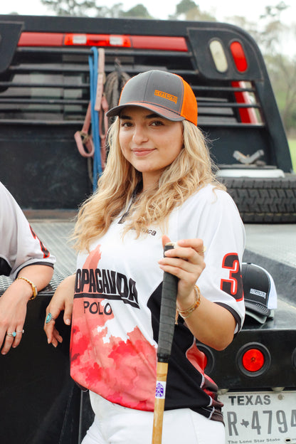 Woman wearing a grey and orange mesh back cap with Texas Polo logo, holding a polo mallet and wearing a jersey.
