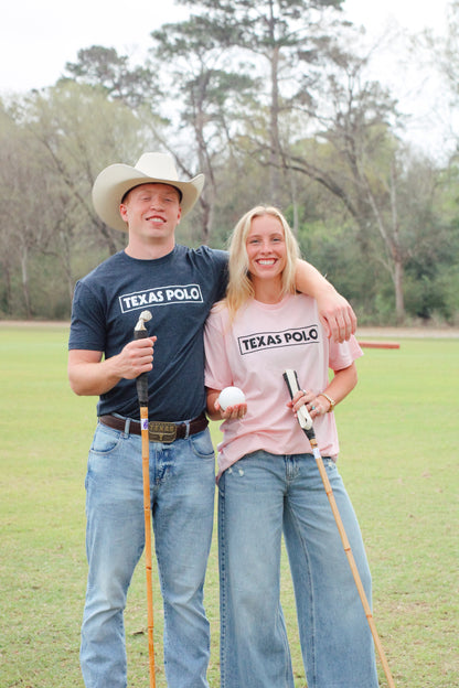 Two people standing on a grassy field holding polo mallets. The boy on the left is wearing a navy t-shirt with a white Texas Polo logo and a tan cowboy hat. The girl on the right is holding a white polo ball and wearing a pink t-shirt with a black Texas Polo logo.