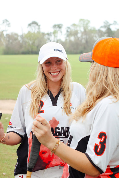 Two women wearing jerseys and holding polo mallets. The woman on the left is wearing a white Texas Polo branded hat, and the woman on the right is wearing an orange and grey Texas Polo mesh back hat.