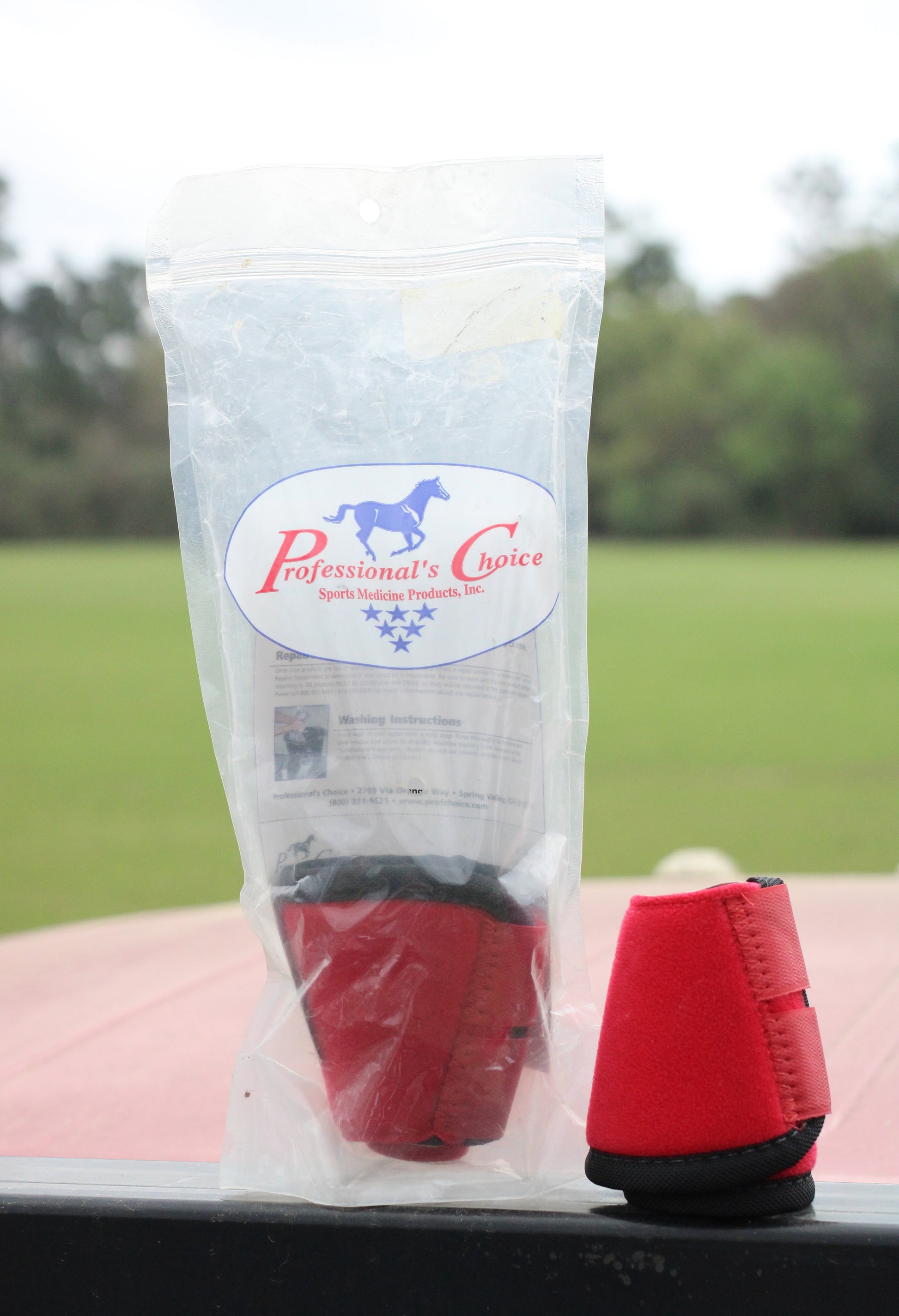 Two red bell boots for horse, with one sitting in a plastic bag and the other sitting next to it.