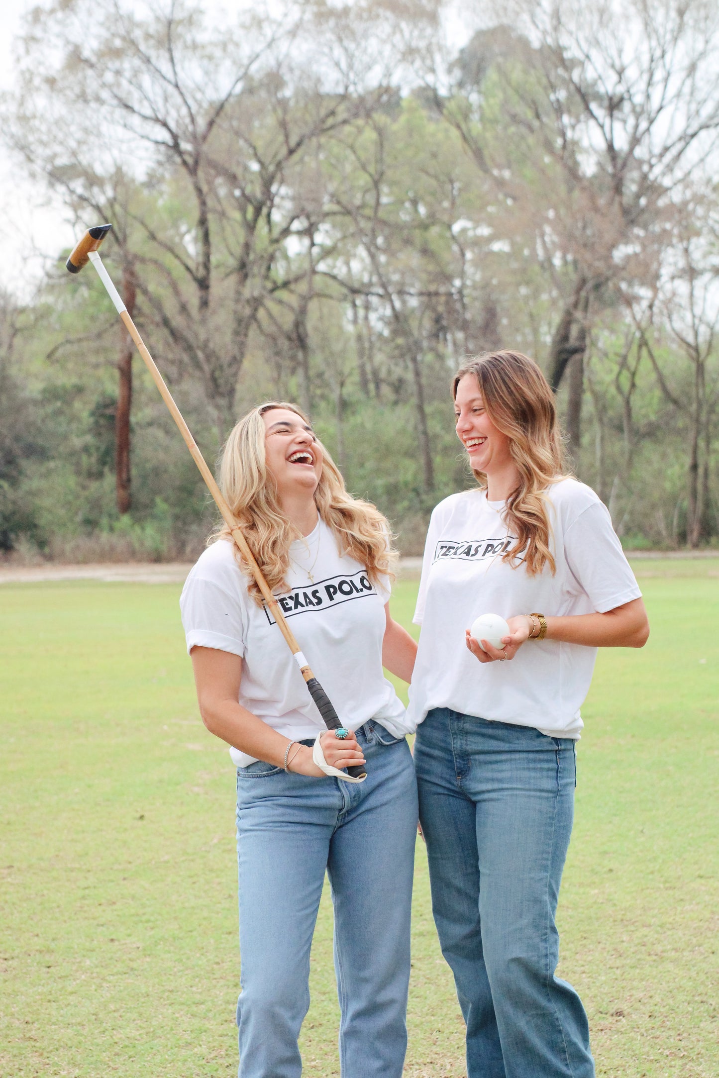 Two women wearing white Texas Polo t-shirts. The woman on the left has a polo mallet over her shoulder, and the woman on the right is holding a polo ball.