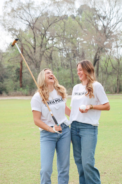 Two women wearing white Texas Polo t-shirts. The woman on the left has a polo mallet over her shoulder, and the woman on the right is holding a polo ball.