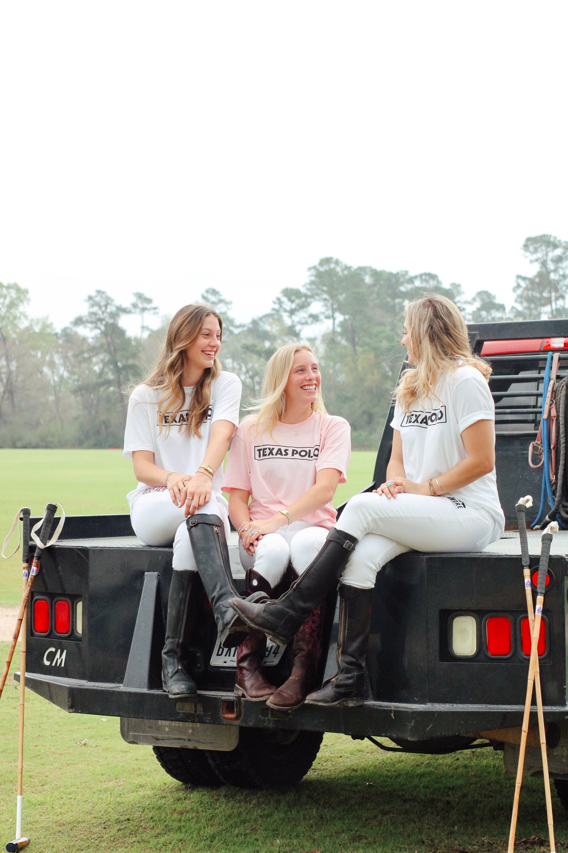 Three women sitting on the back of a truck wearing pink and white Texas Polo branded t-shirts.