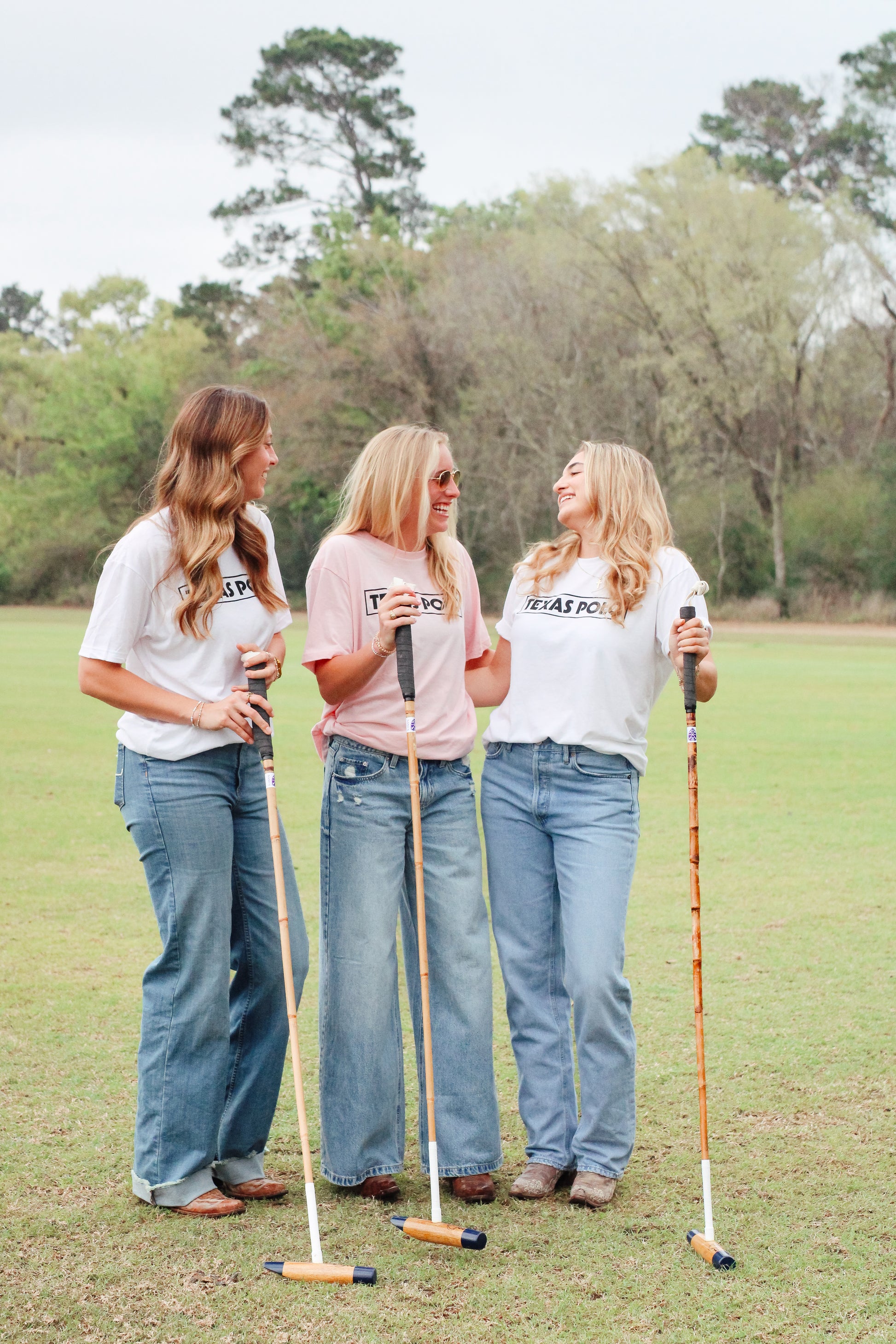 Three women standing on a grassy field holding polo mallets, wearing jeans and Texas Polo t-shirts.