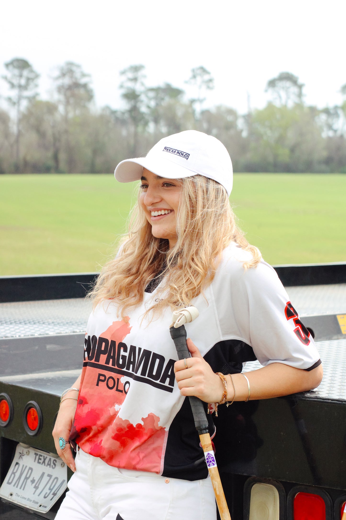 Woman wearing a white Texas Polo branded cap, leaning against a truck bed, wearing a jersey and holding a polo mallet.