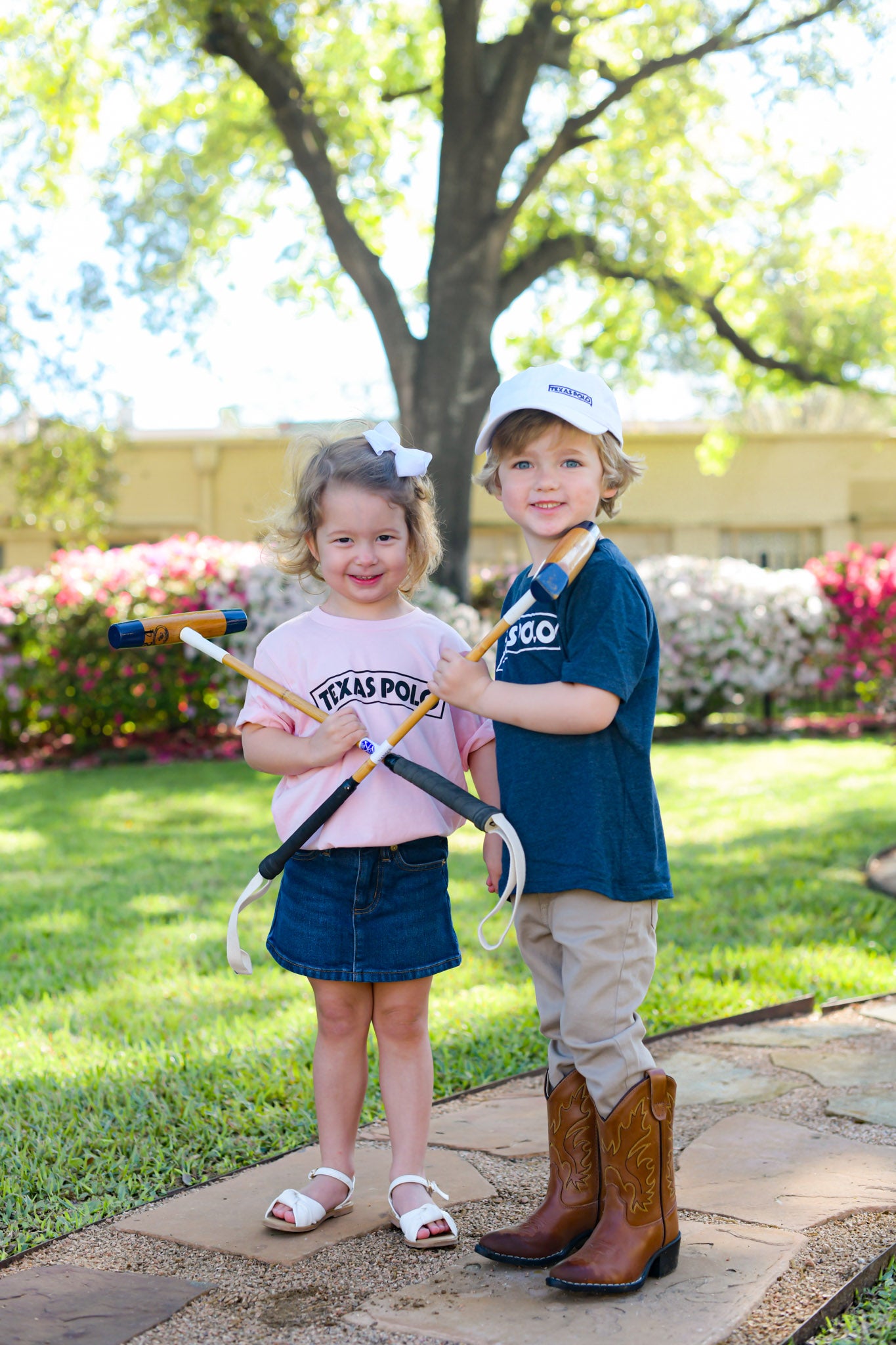 Two children wearing Texas Polo t-shirts in pink and navy and a white Texas Polo hat. They are each holding a foot mallet crossed in front of them.