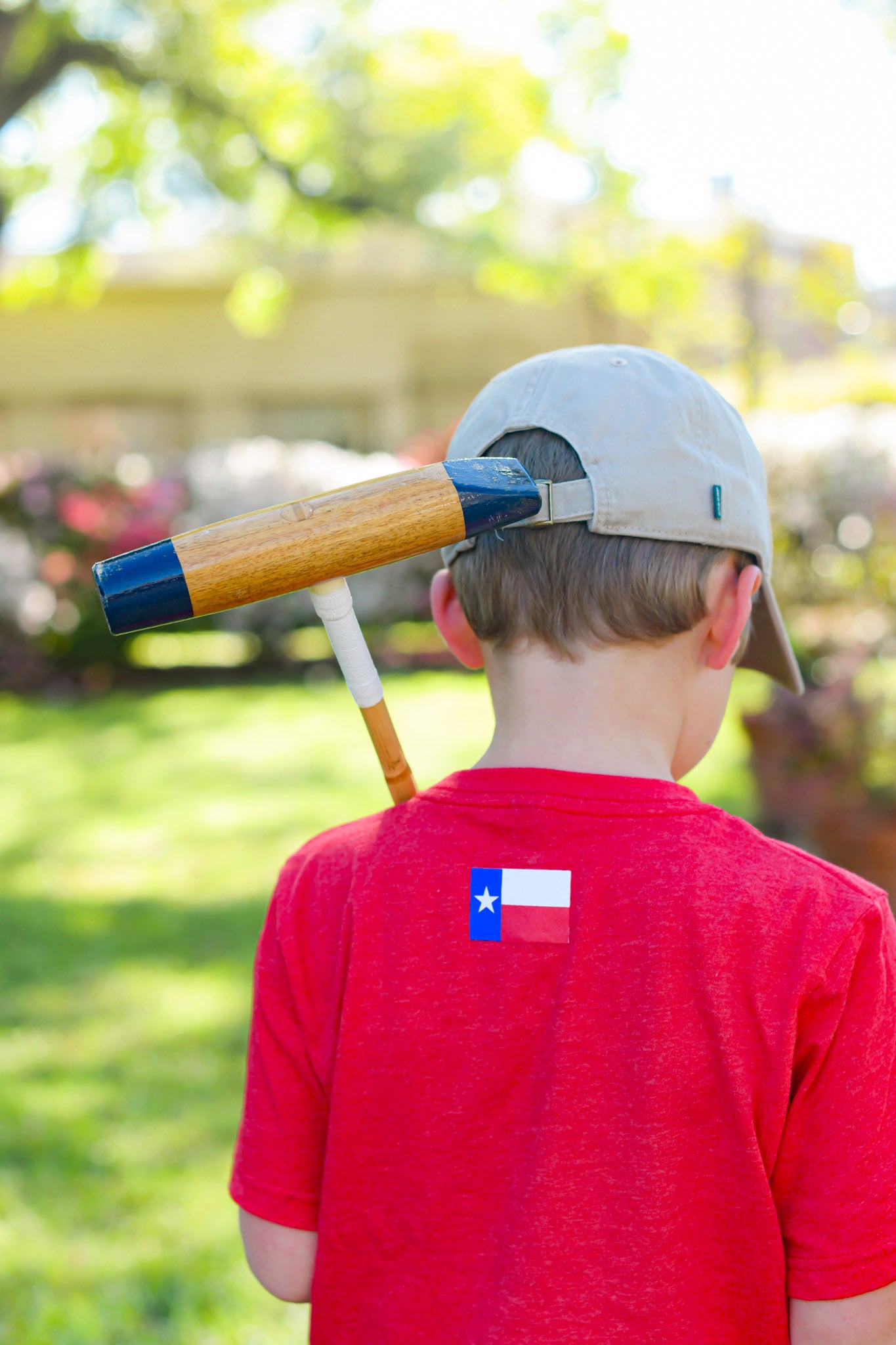 Child holding a polo mallet over his shoulder, wearing a tan cap and a red Texas Polo t-shirt, showing the Texas flag graphic on the back.