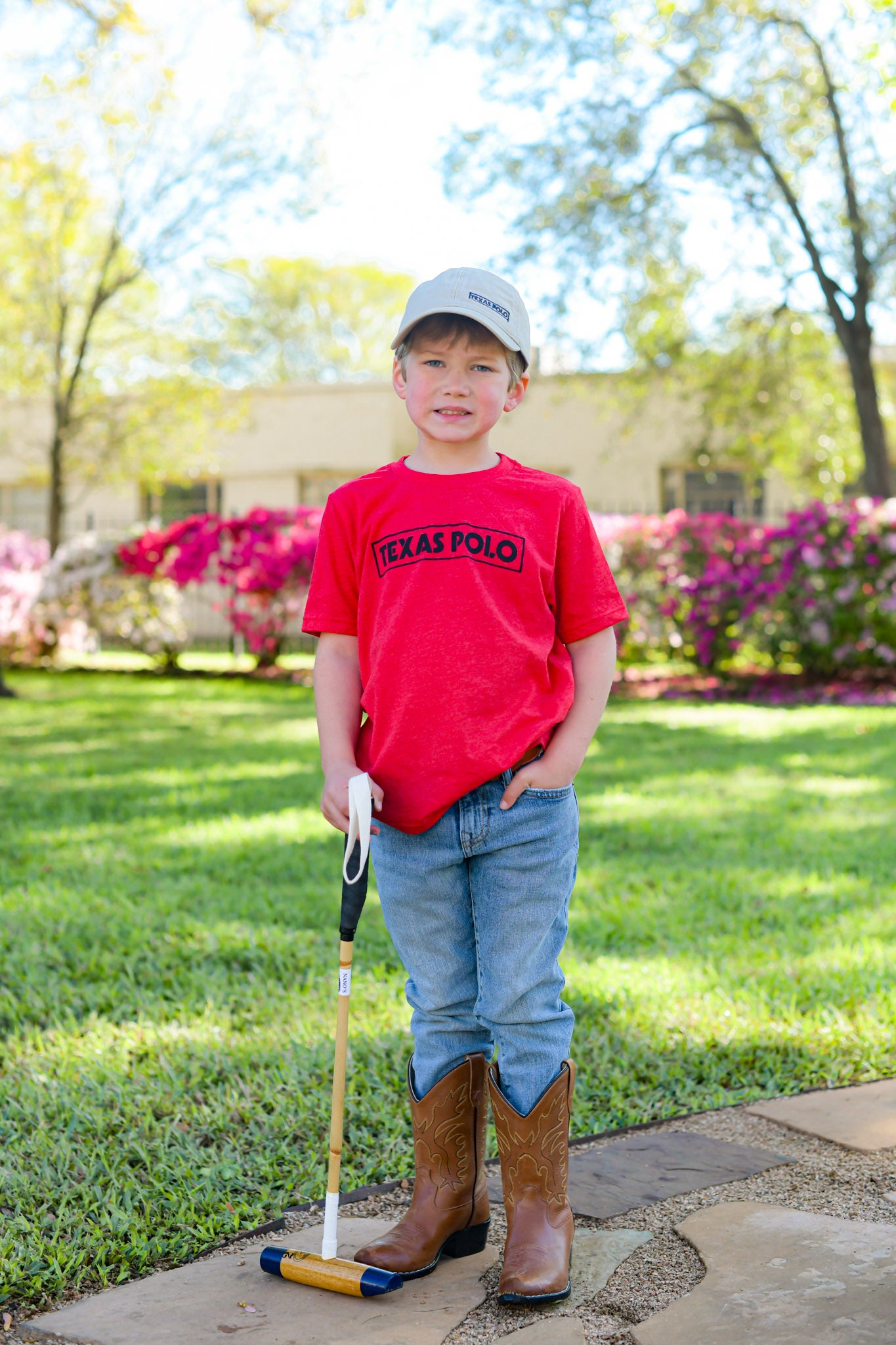 Child wearing a red t-shirt with black Texas Polo logo and tan Texas Polo branded hat, holding a mallet.