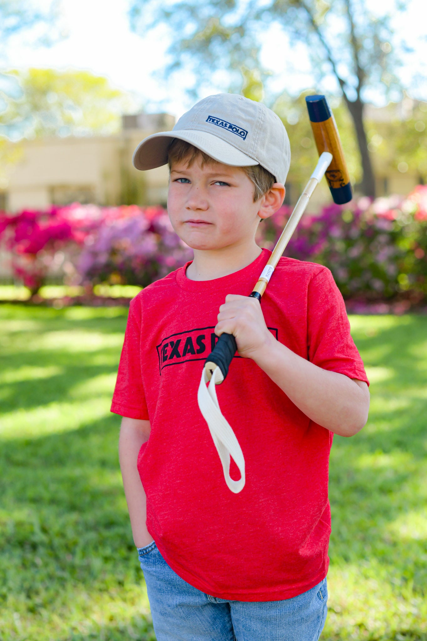 Child wearing a red Texas Polo t-shirt and tan Texas Polo cap, with a foot mallet over his shoulder.