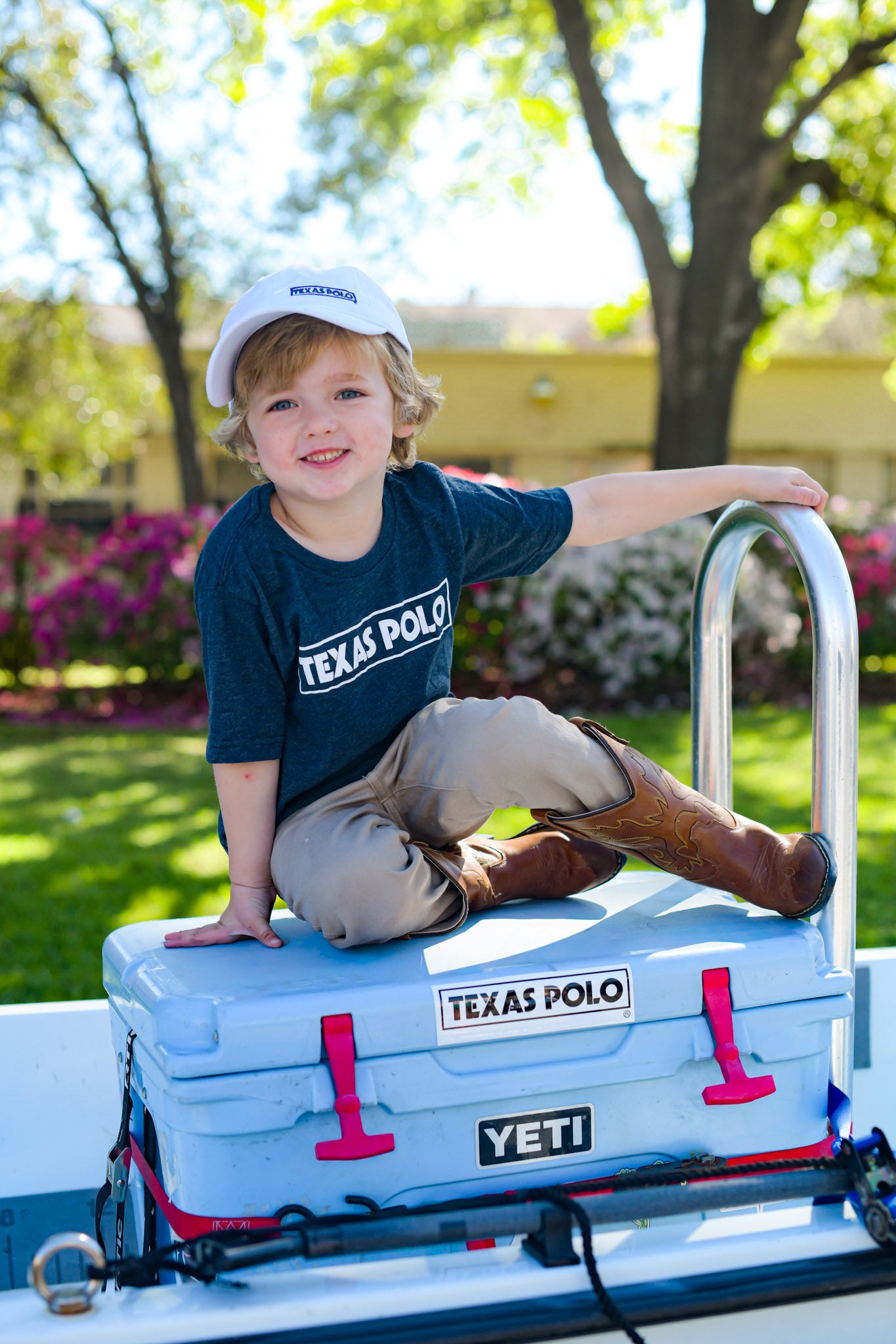 Child sitting on a YETI cooler wearing a white Texas Polo branded cap and a navy t-shirt with a white Texas Polo logo.