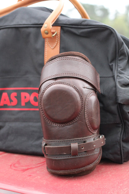 Front view of brown leather knee guard leaning against a black travel bag.