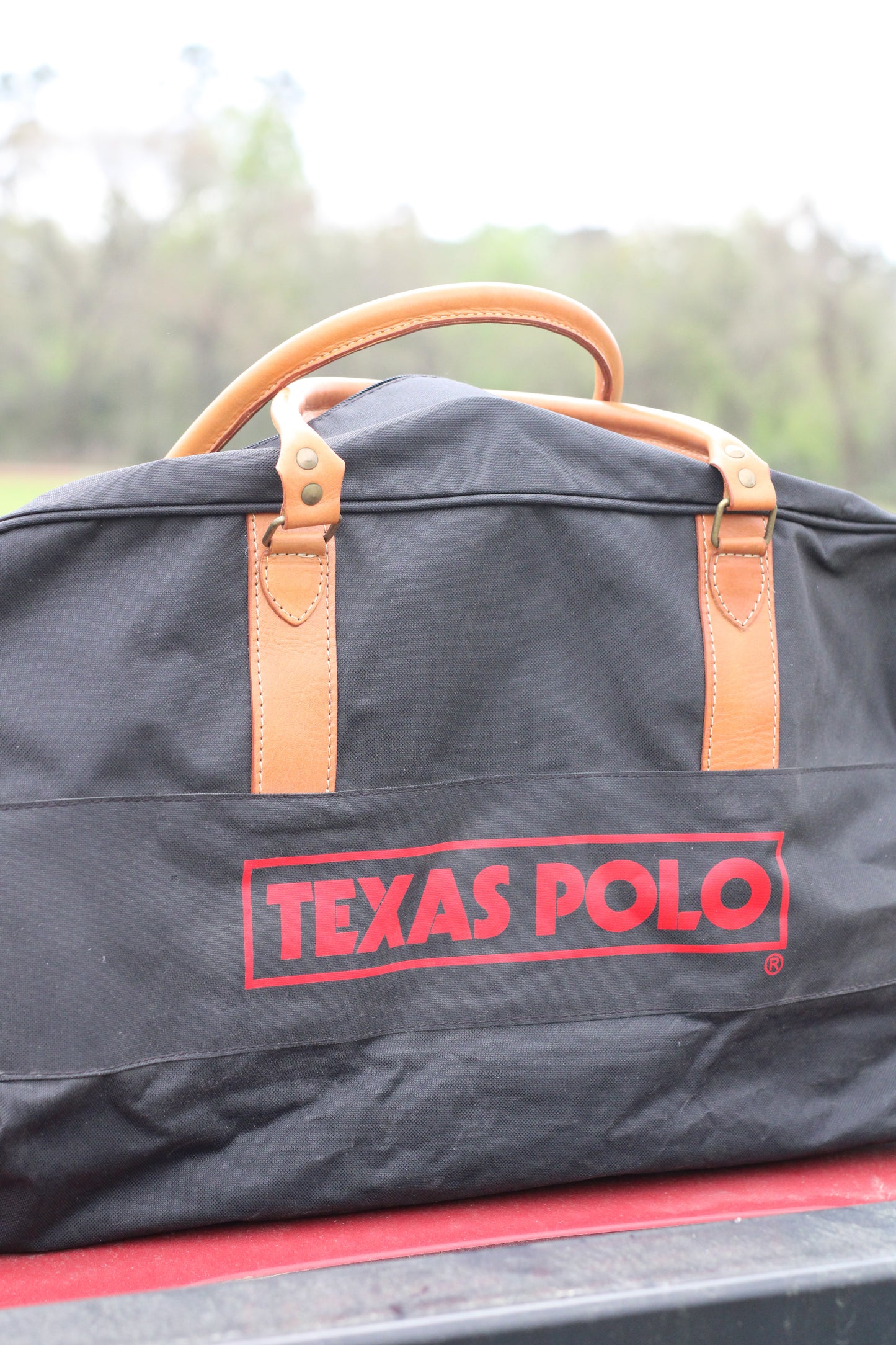 Side view of black travel bag with red Texas Polo logo and tan leather handles.