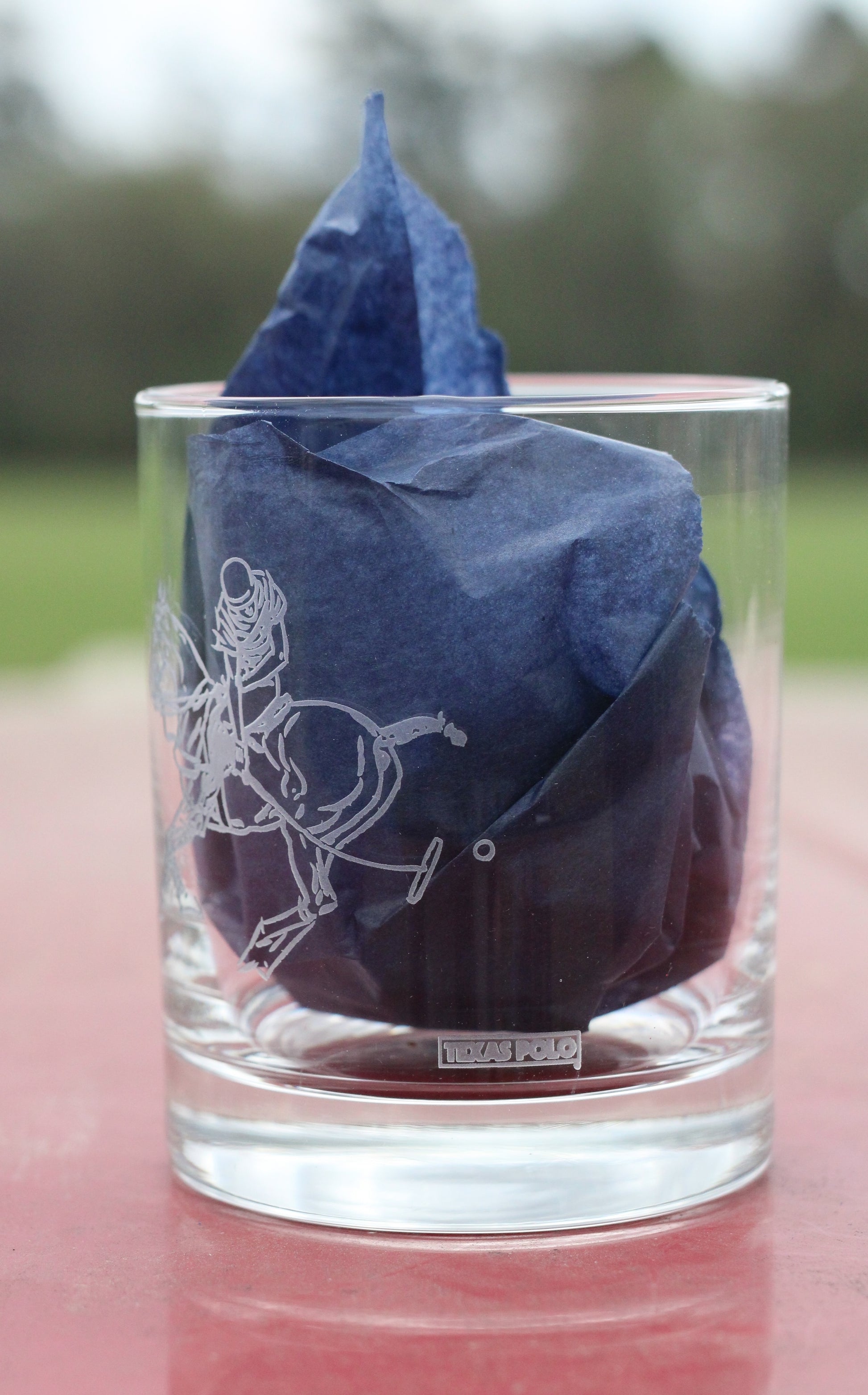 Side view of whiskey glass with polo graphic and Texas Polo logo, with navy tissue paper inside.